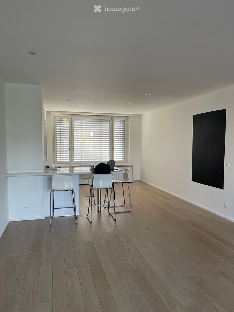 Open-plan room with light wooden flooring, white walls, and a large window with blinds. Features a white bar counter, four white stools, and a small table with a screen on the wall.