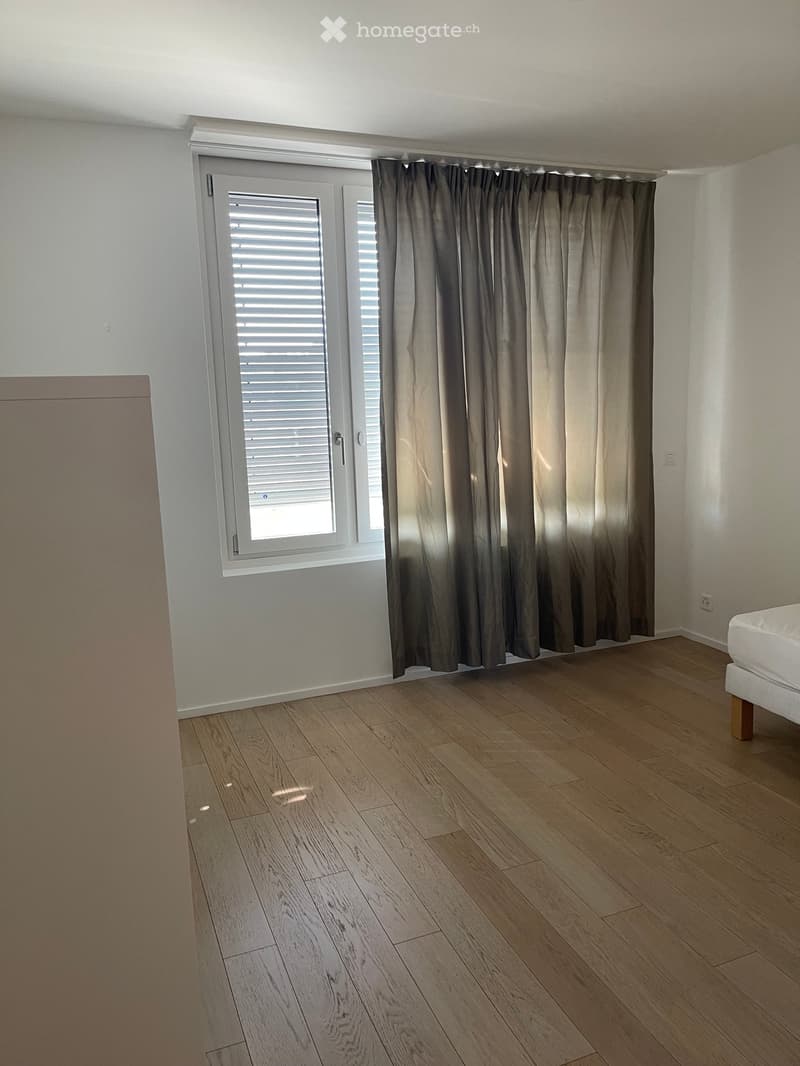 Empty bedroom with wooden flooring, white walls, large window with blinds and curtains, and a partially visible bed frame on the right side.