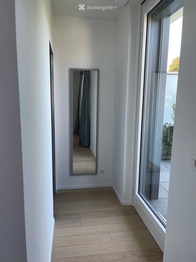 Empty hallway with white walls, wooden flooring, large mirror, and large window with view to outdoor area.