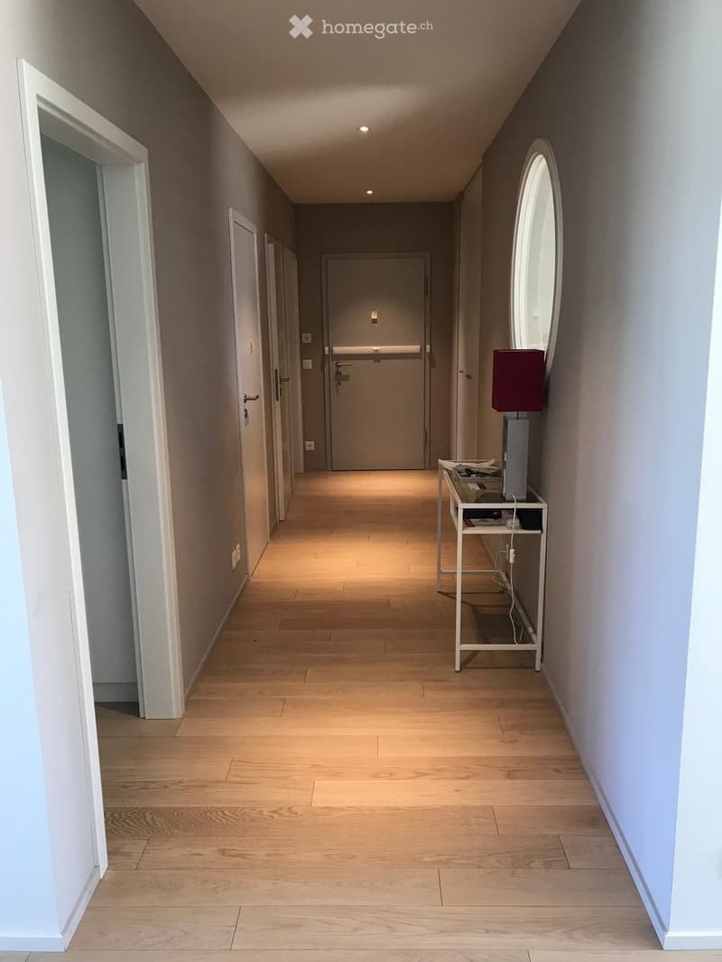 A modern hallway with light wooden flooring, white walls, and several doors leading to different rooms. A small table with a lamp is placed against the right wall.