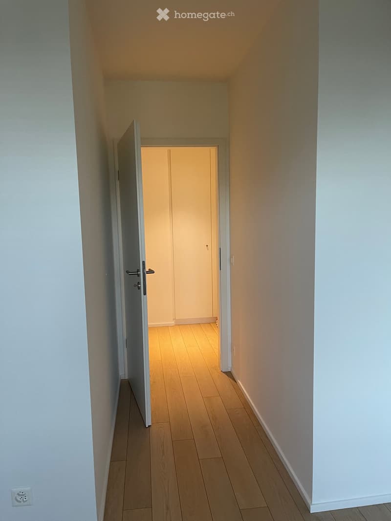 A narrow hallway with light wooden flooring and white walls, featuring an open door leading to another room.