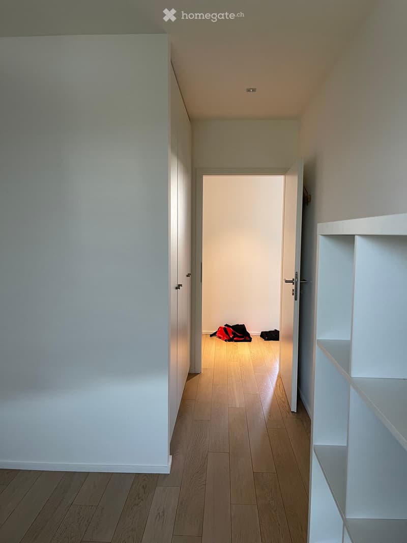 A narrow hallway with light wooden flooring and white walls, leading to an open doorway with a glimpse of another room containing a few bags on the floor.