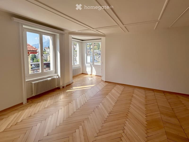 Empty room with wooden herringbone flooring, white walls, large windows with views, and radiators along the walls.