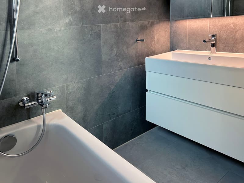 Modern bathroom with a bathtub, sink, and chrome faucets. Grey tiled walls and light grey floor.