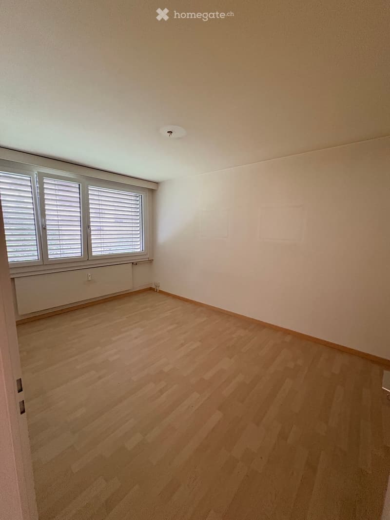 An empty room with wooden floors, white walls, a window with blinds, and a white radiator. There is a door on the left side of the room.