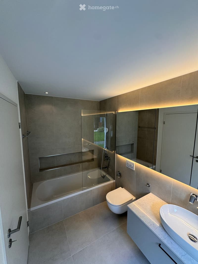 Modern bathroom with a bathtub, toilet, sink, and large mirror. Features include a glass-enclosed shower area and built-in lighting along the walls.