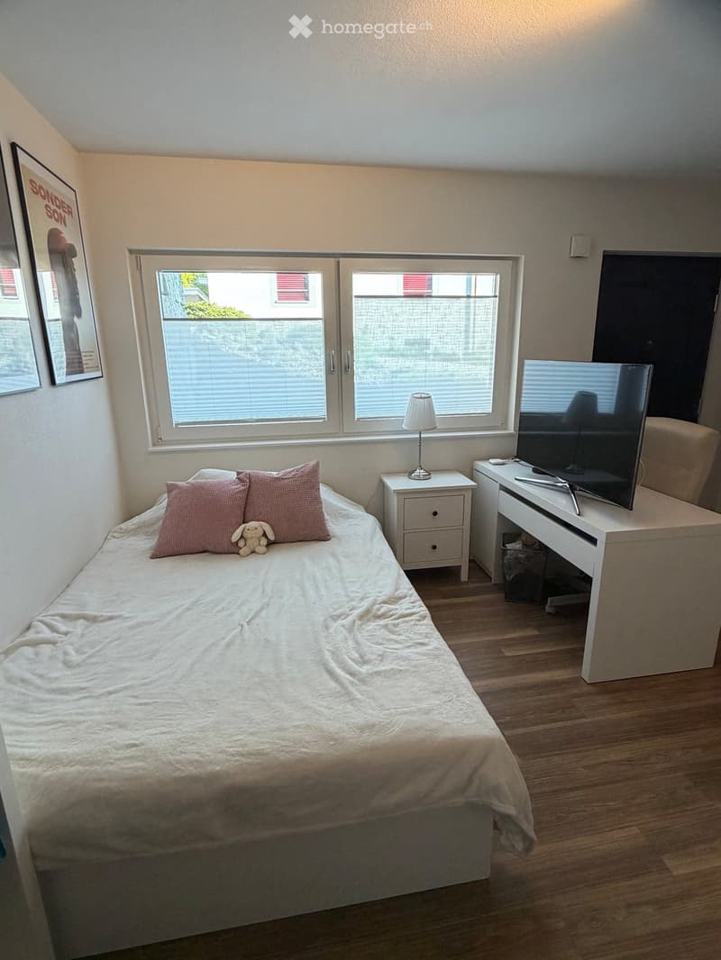 Cozy bedroom with a single bed, nightstand, table lamp, TV stand with a flat-screen TV, and a small stool. The room features wooden flooring and large windows with blinds.