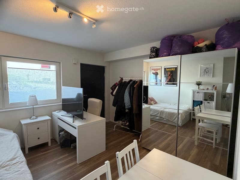 A compact, well-lit bedroom with a bed, desk, chair, table, lamp, mirror, wardrobe, and screen. The room features wooden flooring and large windows.