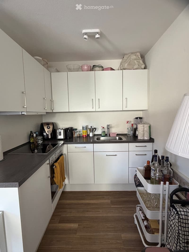 Modern kitchen with white cabinets, dark countertops, and wooden flooring. Visible appliances include an oven, toaster, and blender. A cart with bottles and a towel is placed near the corner.