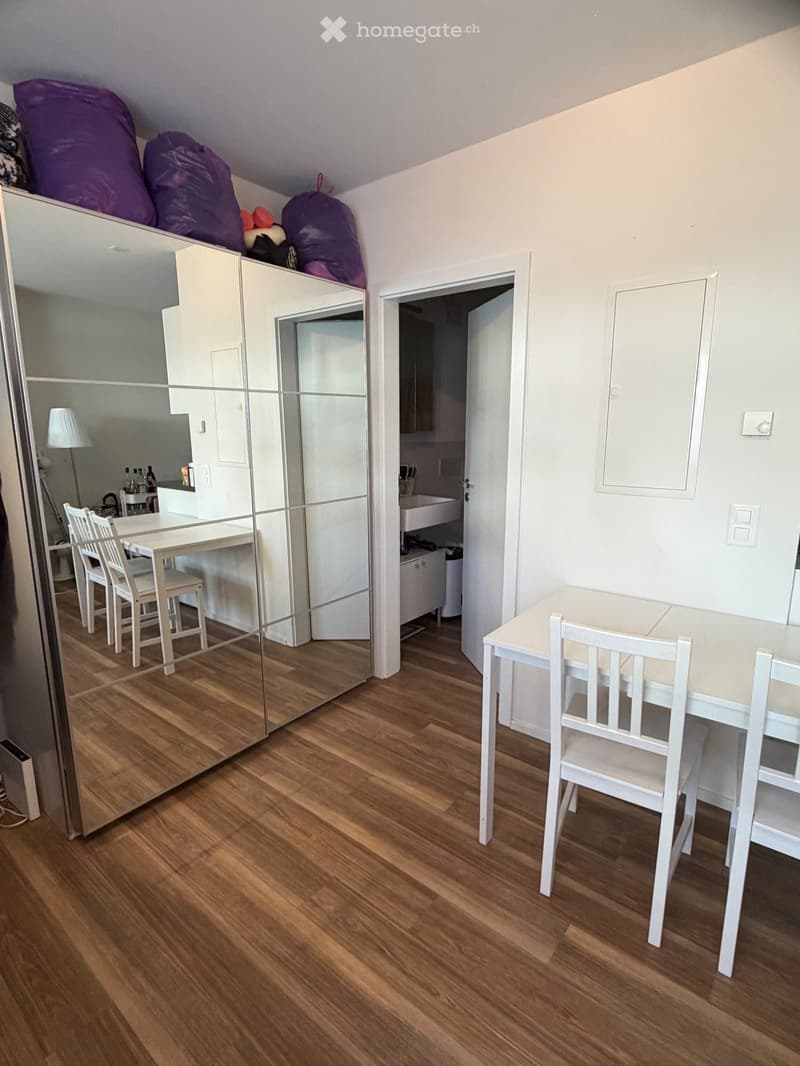 Bright, modern room with wooden flooring, a large mirror, a wardrobe, a small table with chairs, and an open doorway leading to a bathroom.