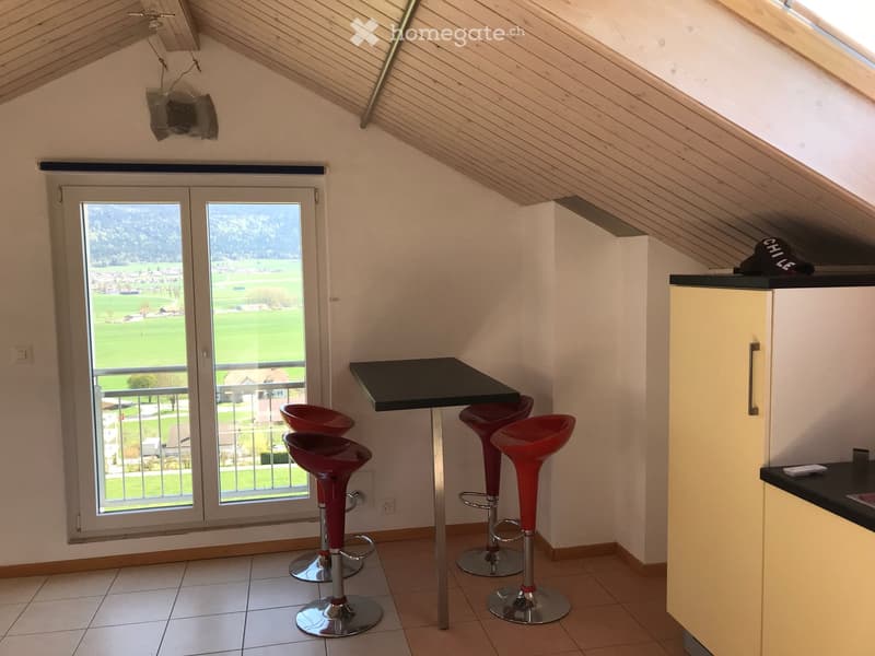 Bright room with a slanted wooden ceiling, large window offering a scenic view, a black table with four red stools, and a light-colored cabinet.