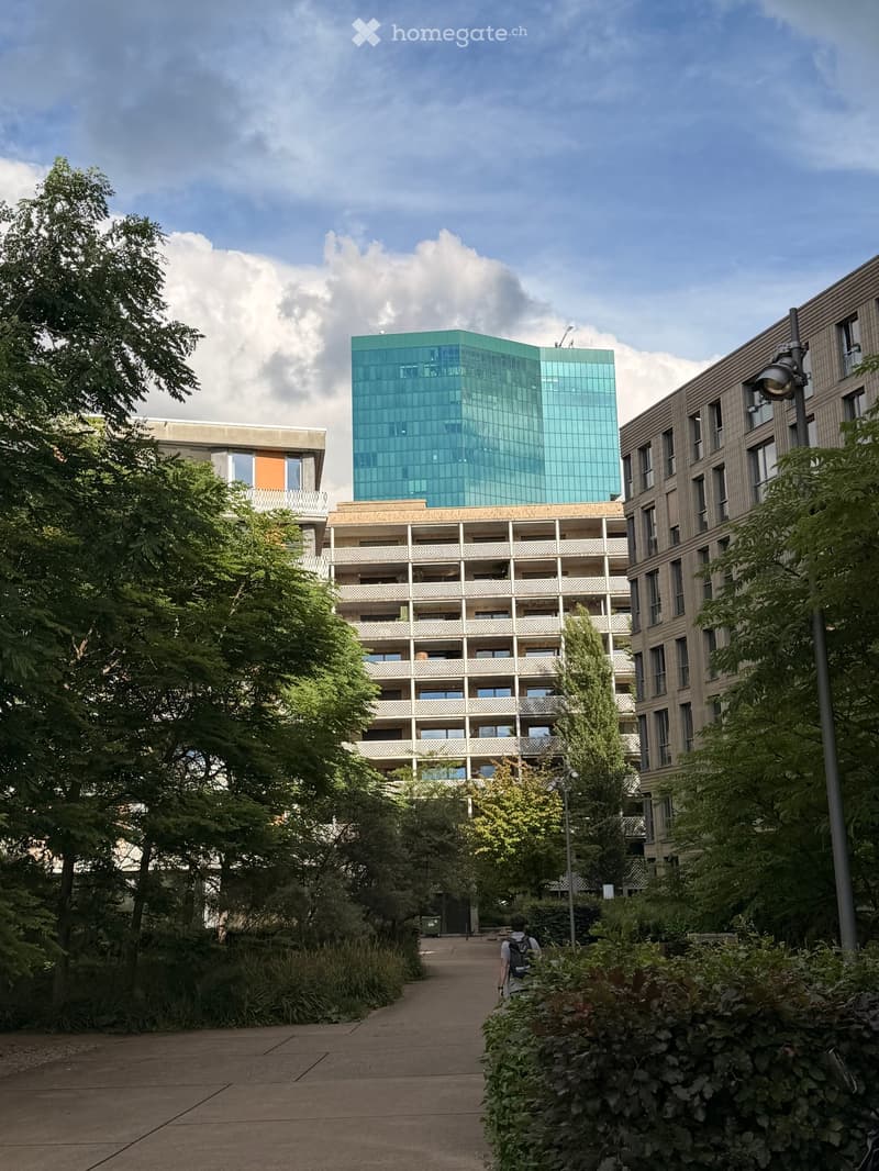 Modern multi-story building complex with glass and concrete structures, surrounded by greenery and pathways.
