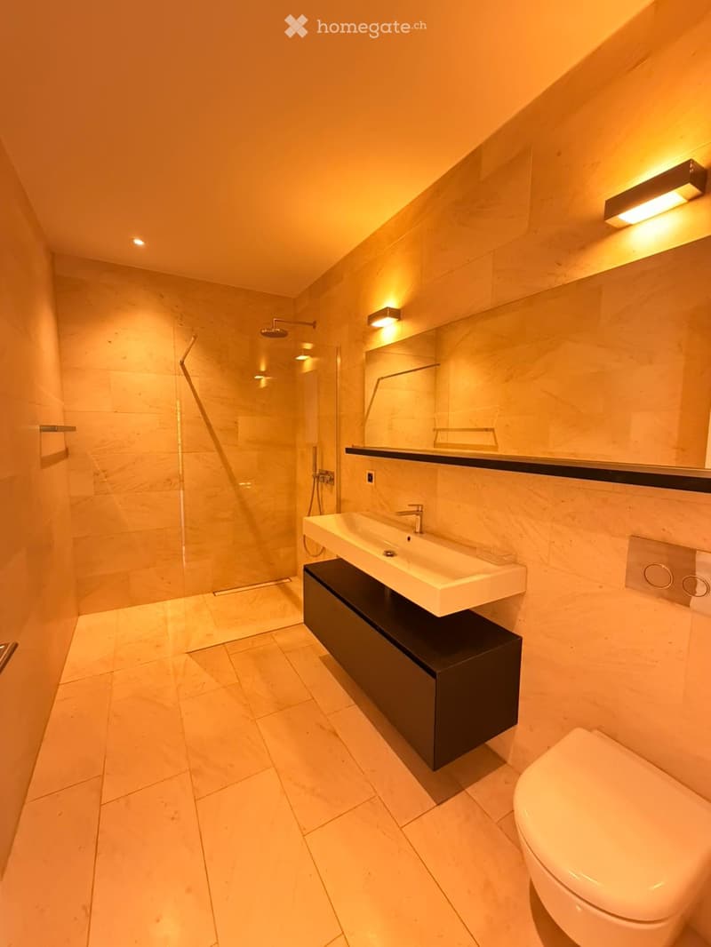 Modern bathroom with a sleek sink, toilet, large mirror, and glass-enclosed shower. Warm lighting and marble tiles.