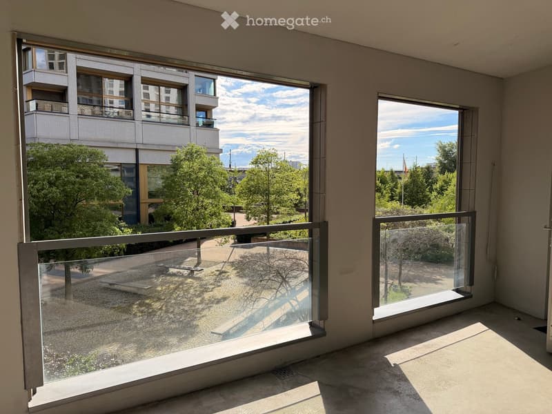 Empty room with large windows and a balcony overlooking a modern building and greenery.