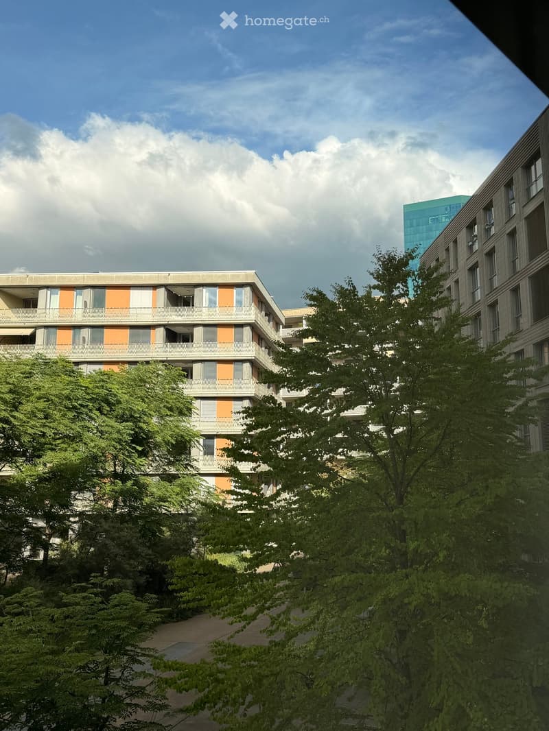 Modern multi-story residential building with balconies and orange accents, surrounded by lush green trees and a clear blue sky.