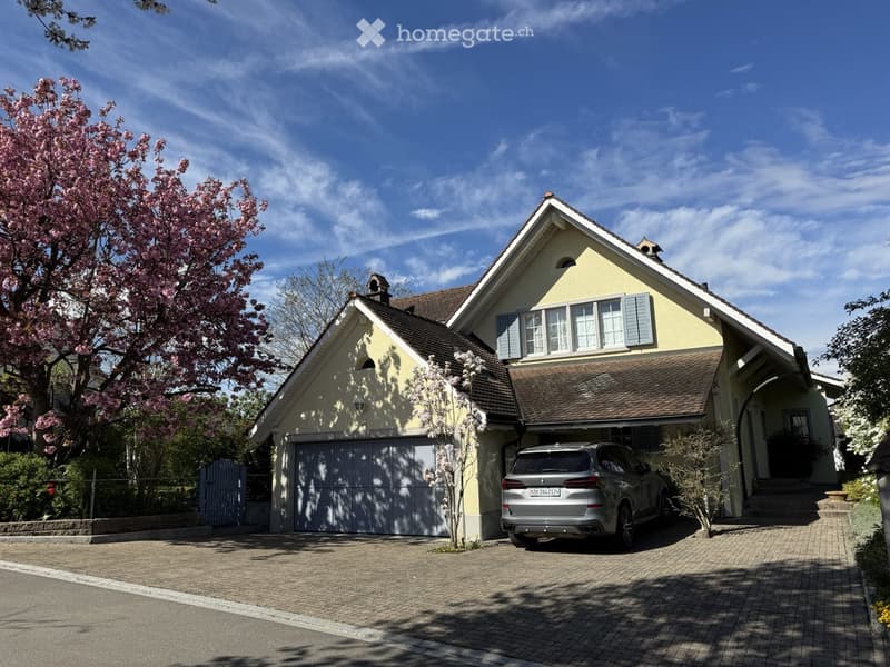 House. Two-story yellow house with a brown tiled roof, garage, and parked car in front. The house features multiple windows with shutters and is surrounded by trees and shrubs.
