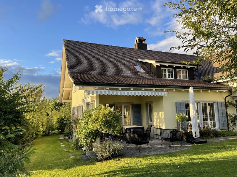 House. Two-story yellow house with a dark roof, multiple windows, and a covered porch. The garden features a patio with outdoor furniture, including a table and chairs, surrounded by greenery and a well-maintained lawn.