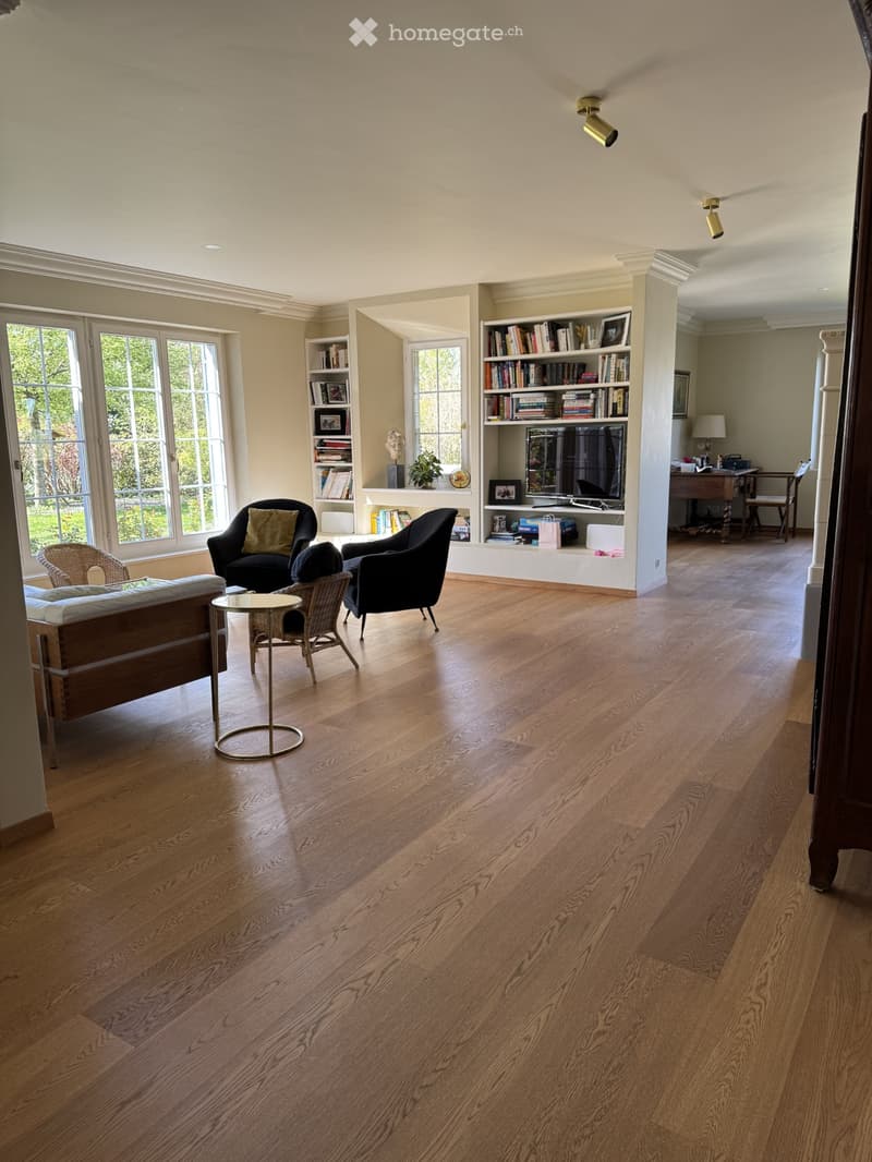 Living Room. Spacious living room with hardwood floors, large windows, a sofa, chairs, a small round table, a bookshelf, and a television.