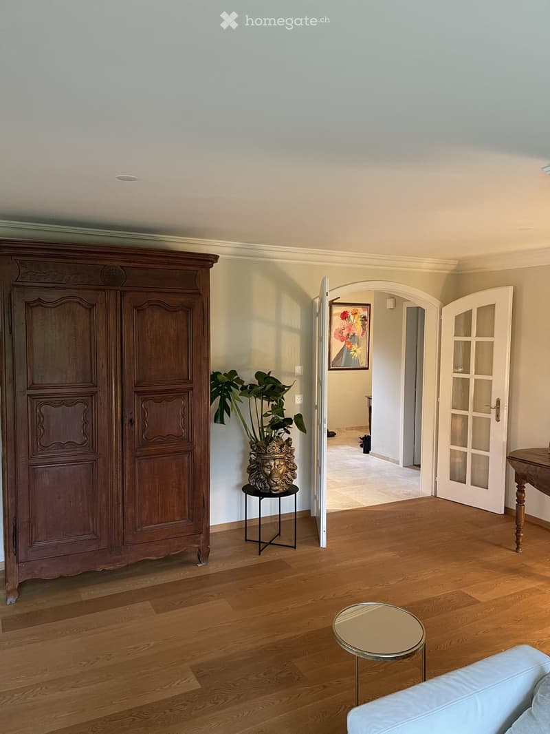 Furnished Room. Spacious hallway with wooden flooring, a large wooden cabinet, a small round table, a plant on a stand, and a white sofa visible in the foreground.