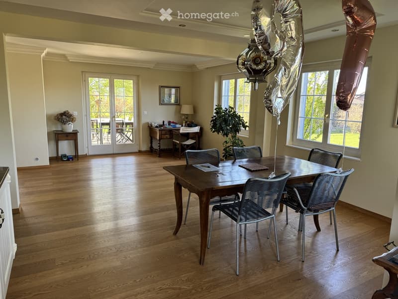 Furnished Room. Dining room with wooden floor, large windows, dining table with chairs, desk with chair, lamp, vase with flowers, and plant.