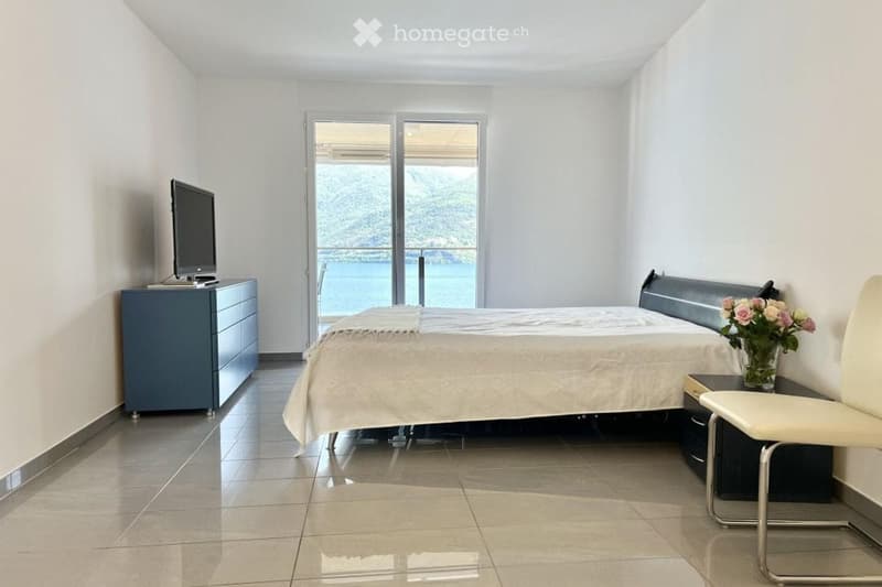 a neatly arranged bedroom with white tiled floor, a large bed, bedside table, flower vase, and a cabinet. The sliding glass door leads to a balcony overlooking the sea.