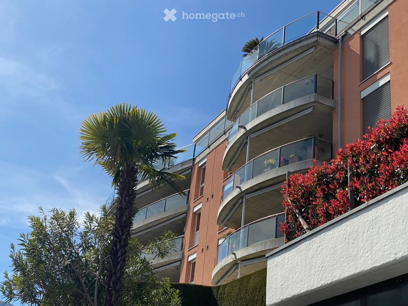 modern apartment building, several floors, balconies with glass railings, greenery, blue sky