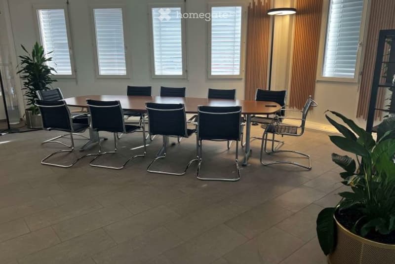 Furnished Room. A conference room with a large wooden table surrounded by black chairs, large windows with blinds, and potted plants.