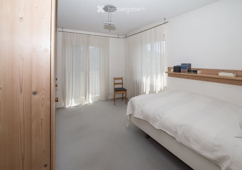 a room with white walls, gray carpet, wooden chair, bed, books on the shelf, curtains on the windows