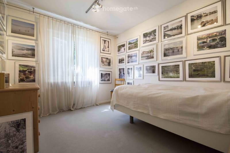 Bedroom with wall covered in picture frames, white bed with white sheets, white floor, wooden dresser, white curtains.