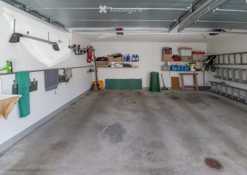 Garage with clean concrete flooring, white walls, and various storage racks with items. Ladders, tools, and storage boxes are neatly organized on shelves and hooks. A green trash bin and a bench are visible. Empty space without any vehicles.