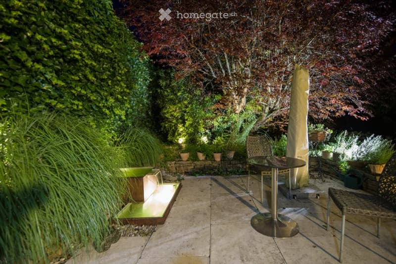 evening scene, outdoor dining table, chairs, water feature, surrounded by greenery, ambient lighting