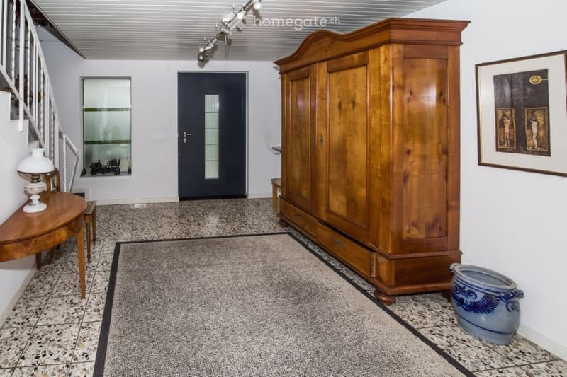 Tiled flooring, white walls, staircase, arched entrance door, wooden cabinet, table with lamp, picture frame