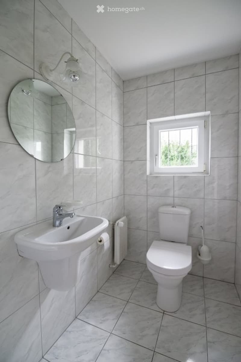 Bathroom: white tiled floor and walls, white sink, toilet, mirror, radiator, window