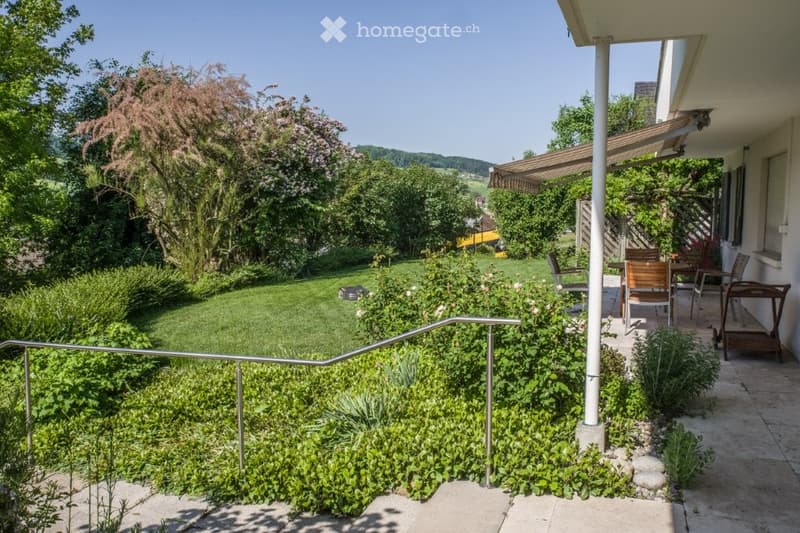 A garden with green grass, plants, wooden chairs, and an awning on a balcony