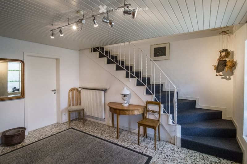 Room features a carpeted floor, white walls, white ceiling with mounted track lights, a wooden staircase with white railing, a lamp on a wooden side table, two wooden chairs, a mirror, and a radiator against the wall.