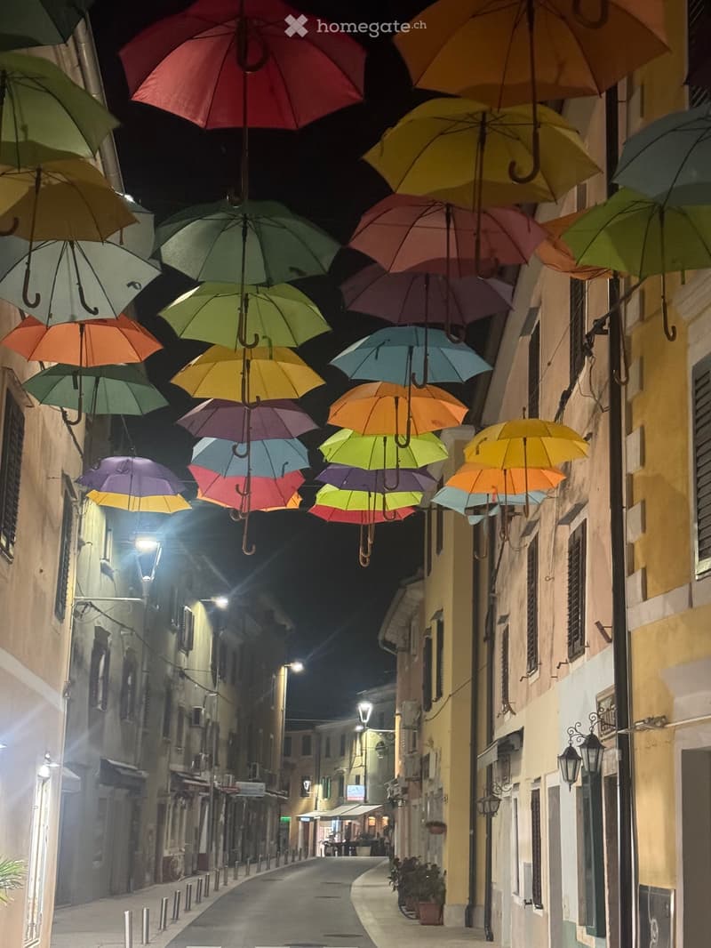 A narrow street lined with colorful hanging umbrellas, illuminated by streetlights, with buildings on both sides.