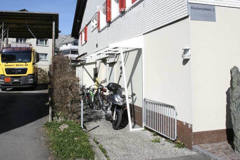 Building. A two-story building with white walls and red shutters, featuring a covered entrance area with bicycles and a motorcycle parked nearby. A truck is visible in the background, and the building has a sign indicating "Klostermühlestr. 3".