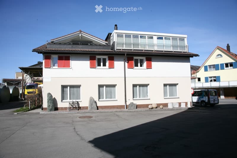 Building. Two-story building with white walls, red shutters, and a balcony on the top floor. The building has multiple windows and a small entrance area with stone decorations.