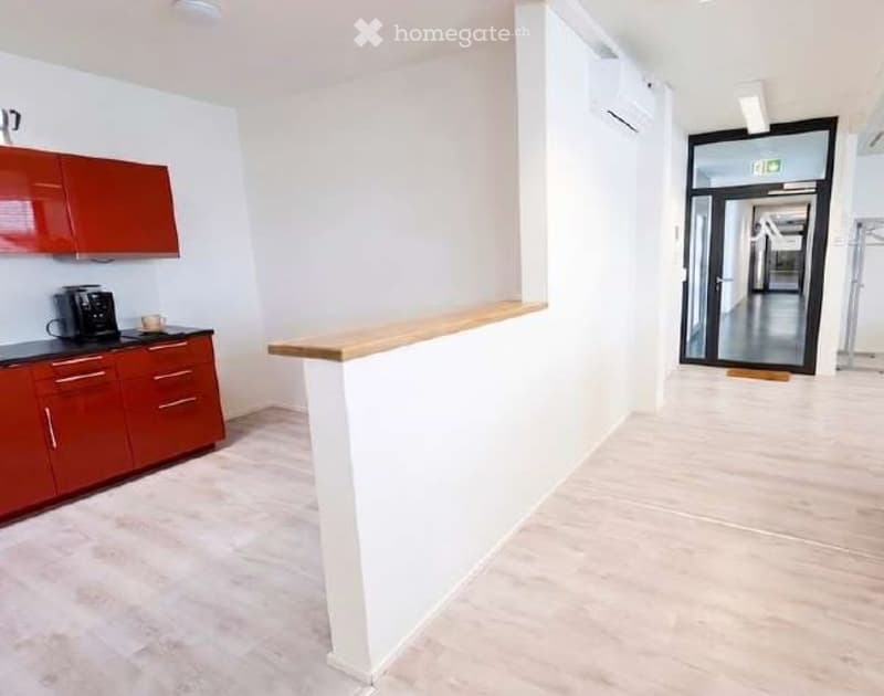 Furnished Room. A modern, minimalist reception area with a wooden counter, red cabinets, and a coffee maker on the counter. The space features white walls, light-colored flooring, and a glass door leading to a hallway.