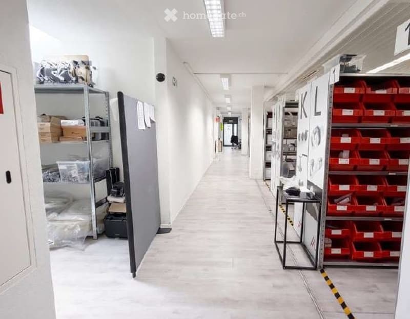 Corridor / Hallway. Clean, well-lit hallway with white walls, tiled floor, and storage shelves on both sides. The hallway features a grey partition, a small table, and red storage bins.
