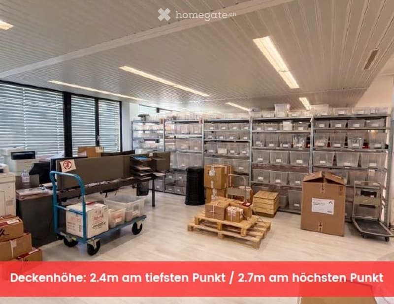 Furnished Room. A spacious, well-lit storage or office area with metal shelving units filled with storage bins, several pallets with boxes, a rolling cart, and a small desk with a computer monitor. The ceiling has exposed beams and fluorescent lighting.