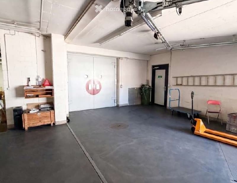 Furnished Room. A spacious, industrial-style room with a concrete floor, white walls, and a large garage door. Visible items include a wooden shelf, a small cabinet, a chair, a pallet jack, and a storage unit.