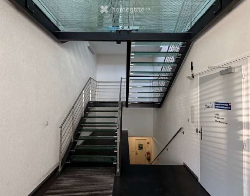 Corridor / Hallway. Modern interior staircase with metal railings and glass ceiling panels, leading to an upper level. The walls are white, and the floor is dark with a textured finish.