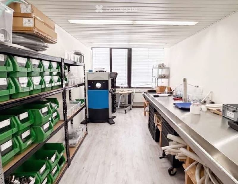 Furnished Room. A well-organized workshop or studio space with white walls, wooden flooring, and large windows. Shelves on the left hold green storage bins and other supplies, while a worktop on the right has tools and a machine. The room is clean and fun