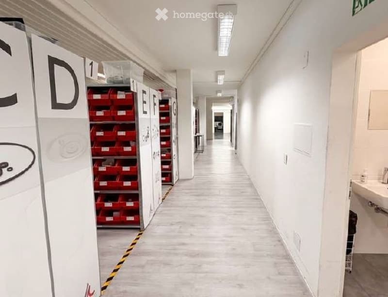 Corridor / Hallway. Clean, modern hallway with white walls, light-colored tiled floor, and storage units with red bins on the left side. Fluorescent lighting and a visible restroom area on the right.