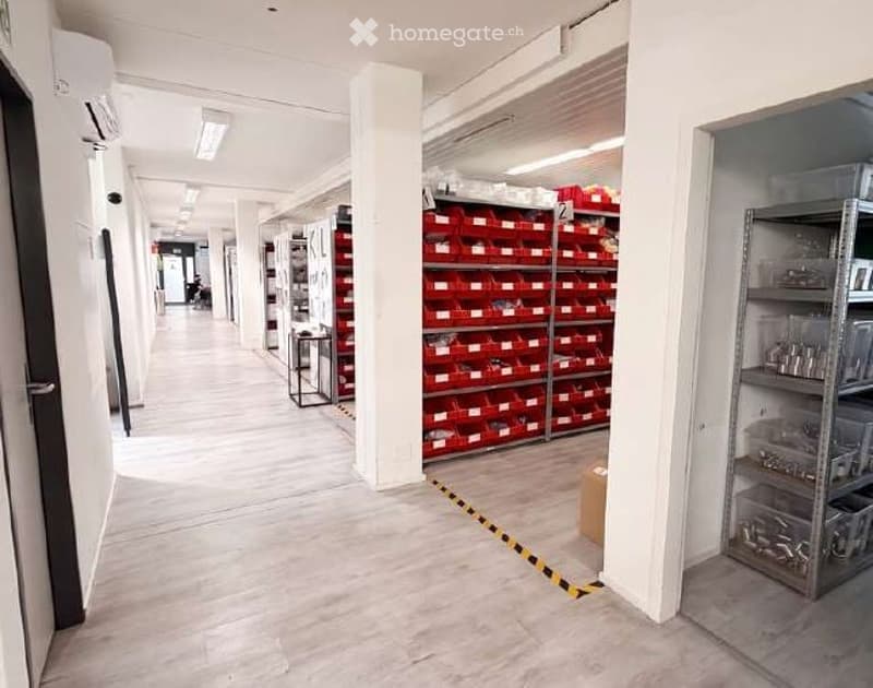 Corridor / Hallway. Clean, well-lit hallway with white walls, tiled floor, and storage shelves on both sides. Red storage bins and metal shelving units are visible.