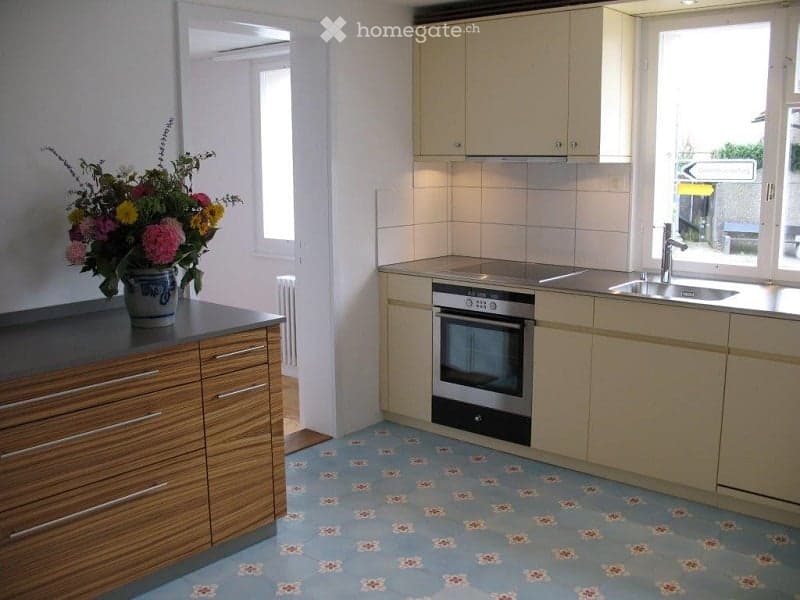 Kitchen. Bright kitchen with light-colored cabinets, stainless steel sink and oven, and a wooden countertop with a decorative flower vase. The floor features a blue and white patterned tile design.