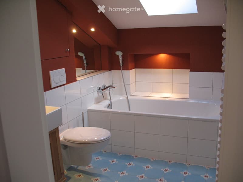 Bathroom. Modern bathroom with a bathtub, toilet, mirror, and showerhead. Features white tiled walls, a blue bath mat with floral patterns, and built-in wooden cabinets.