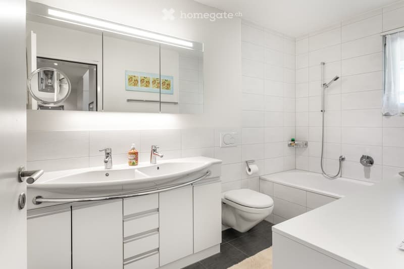Bathroom. Modern bathroom with white tiles, a double sink, a large mirror, a toilet, a bathtub, and a showerhead. Includes a cabinet with drawers and a handrail for accessibility.