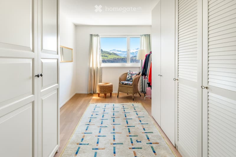 Furnished Room. Bright room with wooden flooring, large window offering a scenic mountain view, a wicker chair with a cushion, a small wicker stool, a patterned rug, a floor lamp, and a wardrobe with hanging clothes.